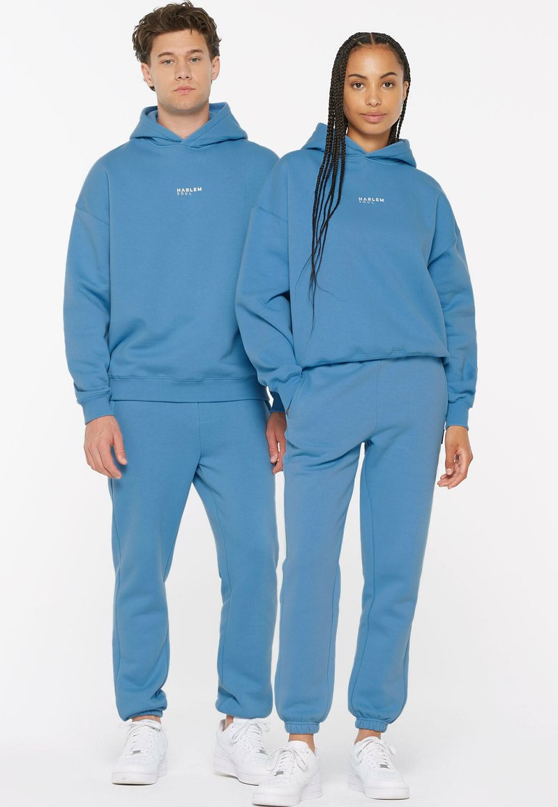 Blaues Baumwoll-Hoodie- und Jogginghosen-Set mit lässiger Passform, gerippten Bündchen und einem kleinen Logo auf der Vorderseite. Weiße Turnschuhe sichtbar.