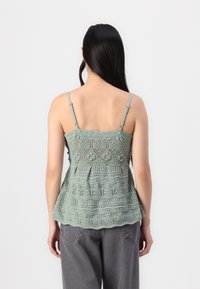 Kvačkan peplum top v mehki zeleni barvi. Značilne so nežne vzorce in prilagodljive naramnice. Teksturna obdelava z razširjenim robom. V kombinaciji s sivimi hlačami.