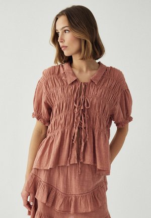 Žena oblečená v textúrovanom krátkokusovom blúzke v farbe prachovej ruže s viazaním a zodpovedajúcej volánovej sukni, pozerajúca sa doľava na jednoduchom pozadí.