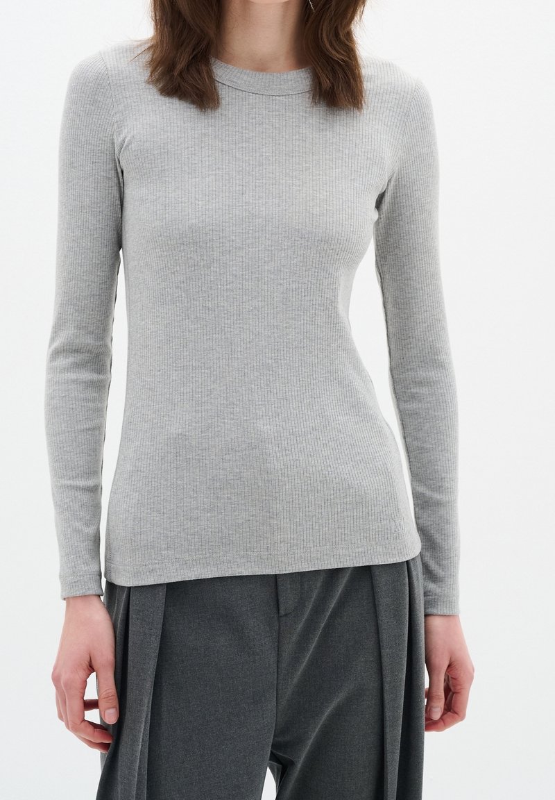 Ljusgrå ribbad långärmad topp med rund halsringning. Tygets struktur är mjuk, med en figurnära silhuett, i kombination med mörkgrå veckade byxor.