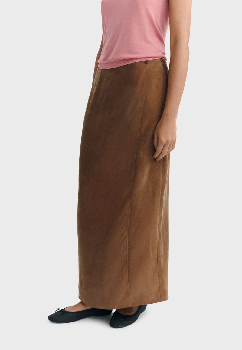 Femme portant une longue jupe en daim marron, un haut rose à manches courtes et des ballerines noires, tenant debout devant un fond uni.