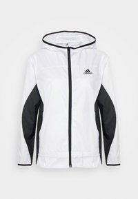 Bílá bunda s kapucí a černými bočními panely, s celým zipem a logem Adidas na levé straně hrudi. Materiál působí lehce a lesklý.