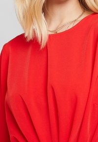 Rød bluse med blød tekstur, med rund halsudskæring og samlede detaljer ved taljen. Lagdelte guldkæder.