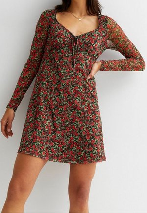 Kvinde iført en langærmet, kort blomsterkjole med røde blomster og grønne blade, stående med den ene hånd på hoften mod en ensfarvet baggrund.