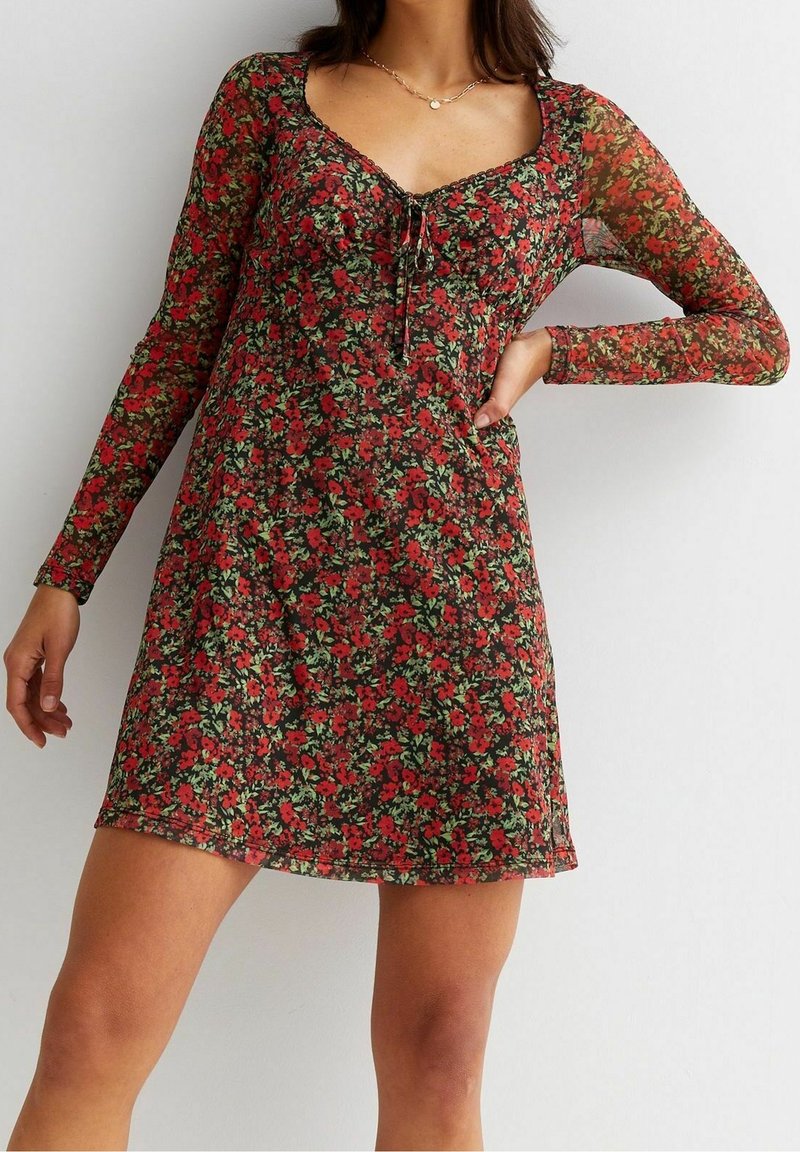 Femme portant une robe courte à manches longues à fleurs rouges et feuilles vertes, debout avec une main sur la hanche devant un fond uni.