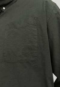 Gros plan d'une chemise en coton vert foncé montrant une poche poitrine et une partie de l'épaule et du col avec un bouton blanc.