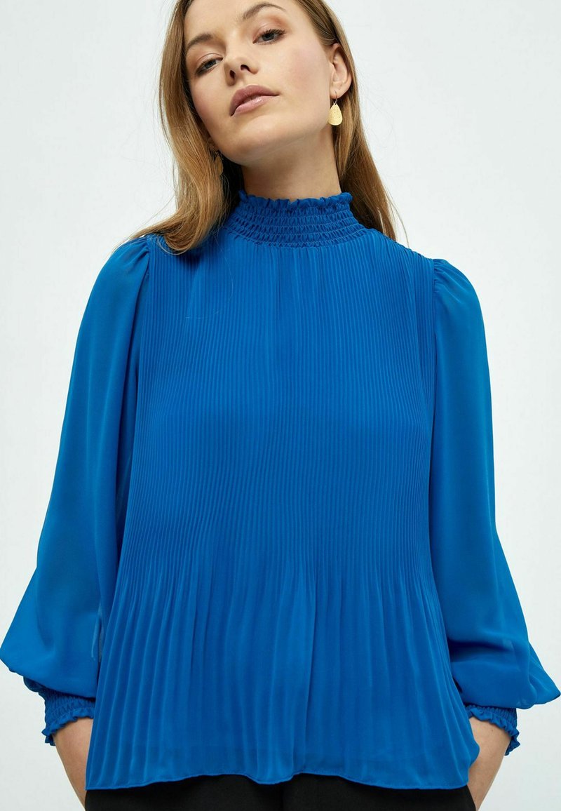 Top blu plettato con un alto collo smock, maniche lunghe a sbuffo e vestibilità rilassata. Tessuto liscio con una leggera lucentezza, dettaglio arricciato sui polsini.