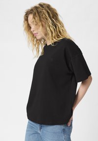 Sort bomulds T-shirt med en afslappet pasform, korte ærmer og et subtilt logo på brystet. Kombinationen er med lyseblå jeans.