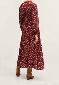Dlouhé vínové šaty s bílými květinovými vzory, elastickým pasem a dlouhými rukávy. Kombinovány s hnědými kotníkovými botami a rozšířenou sukní.