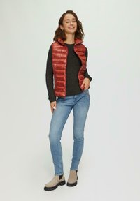 Rode gewatteerde vest met hoge kraag boven een donkere trui, gedragen met lichtblauwe jeans en beige enkellaarsjes met zwarte zolen.