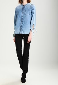 Camicia in denim azzurro chiaro con scollo rotondo, chiusura con bottoni e maniche lunghe con dettagli a fiocco. Presenta una tasca sul petto e cuciture a contrasto.