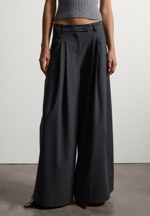 Person trägt dunkelgraue, weit geschnittene plissierte Hose und ein graues, bauchfreies ärmelloses Top, steht auf einem Betonboden.