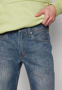 Jean en denim de couleur bleu clair avec un léger effet délavé, doté de cinq poches et de coutures orange, associé à un sweat à capuche vert clair.