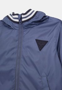 Giacca leggera blu con zip frontale, colletto a coste striato blu navy e bianco, e una patch logo triangolare nera sul petto.