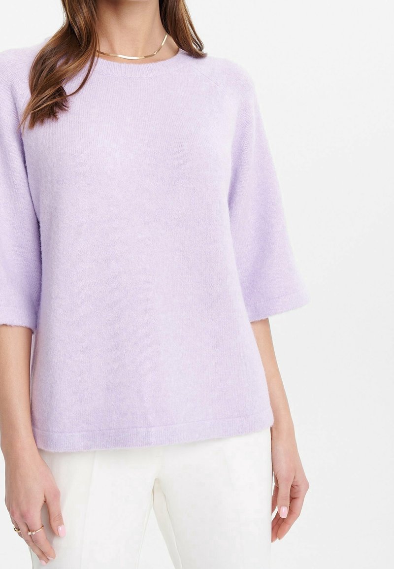 Lila, kurzärmliger Pullover aus weichem Strickmaterial mit rundem Halsausschnitt und lässiger Passform, kombiniert mit weißen Hosen.