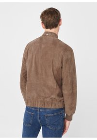 Man met zijn rug naar de camera, gekleed in een brown suede jack met geribbelde manchetten en zoom, in combinatie met blauwe spijkerbroek, tegen een effen achtergrond.