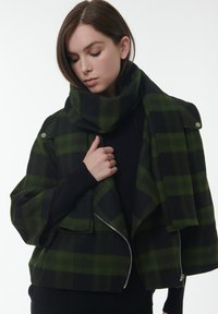 Groene en zwarte geruite jas met een hoge kraag, cropped design, zilveren ritsen en een textiele stof. Het model draagt een top eronder.