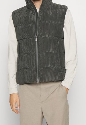 Mężczyzna ubrany w ciemnoszary pikowany kamizelkę z prążkowanego materiału na jasnobeżowej, długorękawowej koszuli i jasnobeżowych spodniach, stoi z jedną ręką w kieszeni.