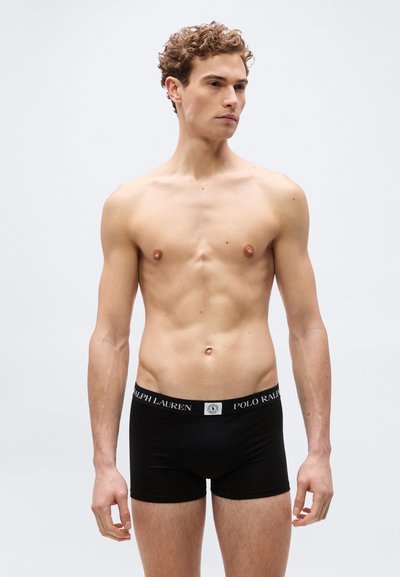 Νεαρός άνδρας με σγουρά μαλλιά στέκεται γυμνόστηθος φορώντας μαύρα εσώρουχα Ralph Lauren boxer briefs, κοιτάζοντας ελαφρώς προς τα δεξιά του, μπροστά σε ένα απλό φόντο.