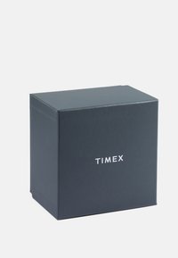 Fekete téglalap alakú doboz, amelyen a "TIMEX" felirat fehér színnel található. Simaságú matt felület, éles szélekkel. Nincsenek látható minták vagy díszítések.