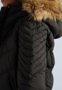 Tuvojoties melnā kvadrātveida ziemas jakas piedurknēm ar dižālo šuvumu un brūnu kažokādu apmalotiem kapuci, kas ir redzams augšpusē.