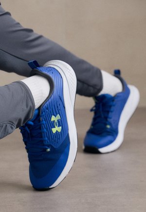 Beine übereinandergeschlagen, weiße Socken, graue Hose und blaue Sportschuhe mit weißen Sohlen auf einem glatten, beigen Boden.