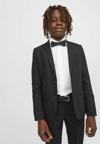 Veste noire à deux boutons, associée à une chemise blanche et un nœud papillon noir. Notable pour sa coupe ajustée et sa texture de tissu lisse.