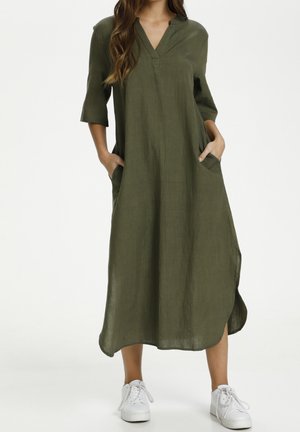 Robe en lin vert olive avec un décolleté en V, des manches trois-quarts et des poches latérales. Présente une coupe ample et un ourlet à la cheville. Portée avec des chaussures blanches.