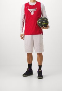 Camiseta de baloncesto sin mangas roja con un gráfico de un toro, combinada con pantalones cortos de malla blancos. Zapatillas negras con acentos morados. Sosteniendo un balón de baloncesto.