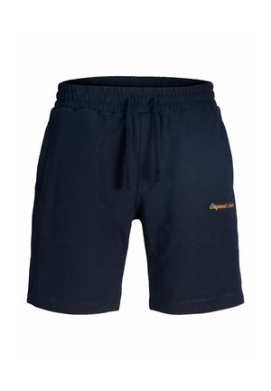 Garçon portant un sweat à capuche bleu marine et un short, avec des chaussettes blanches et des baskets, debout et regardant sur le côté sur un fond uni.