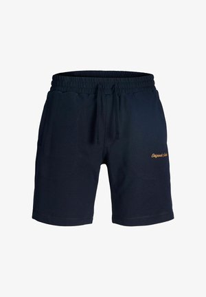 Garçon portant un sweat à capuche bleu marine et un short, avec des chaussettes blanches et des baskets, debout et regardant sur le côté sur un fond uni.