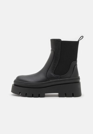 Sort læder Chelsea-støvle med tyk slidstærk sål, elastisk sidepanel og træksnøre på glat grå baggrund.