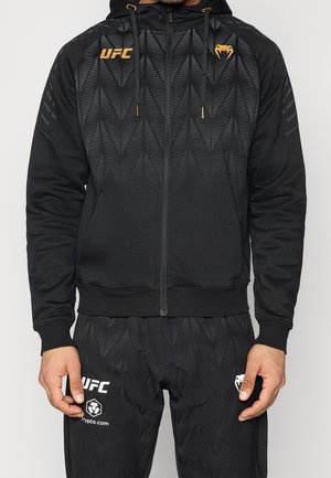 Homme portant un sweat à capuche zippé noir de la marque UFC et un pantalon assorti avec un motif géométrique subtil et des logos dorés et blancs.