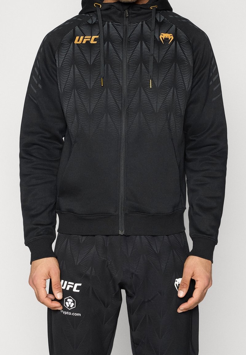 Homme portant un sweat à capuche zippé noir de la marque UFC et un pantalon assorti avec un motif géométrique subtil et des logos dorés et blancs.