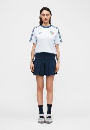 Donna che indossa una t-shirt Adidas azzurro chiaro con stemma dell'Italia, gonna plissettata blu navy, calzini neri e sneakers bianche, in piedi davanti a uno sfondo semplice.