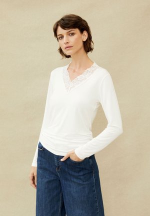 Kvinde med kort brunt hår iført en hvid, langærmet top med blonder i V-udskæring og blå jeans, stående mod en ensfarvet beige baggrund.