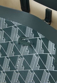 Sac en cuir vert foncé avec un motif géométrique blanc, logo crocodile Lacoste embossé, et poignées en cuir noir sur fond beige.