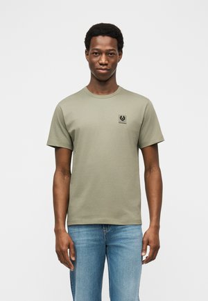 Muž stojící čelem k divákovi, oblečený v světlém olivově zeleném tričku s malým logem na hrudi a světle modrých džínách, na jednobarevném pozadí.