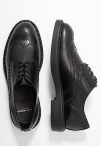 Chaussures brogues en cuir noir présentant des perforations détaillées, un bout arrondi et une semelle basse et robuste. Texture lisse avec des accents cousus.