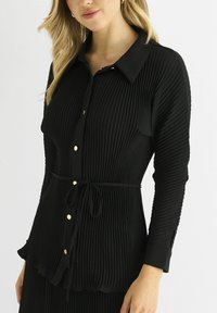 Black, long-sleeve blouse with a textured pleated design, button front, and a tie waist. Features gold-tone buttons and a collar.
