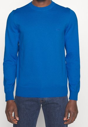 Mann trägt einen einfarbigen blauen Pullover mit langen Ärmeln und einem kleinen "BOSS"-Logo auf der Brust, kombiniert mit dunkelblauen Jeans.