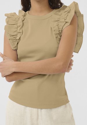 Woman wearing beige sleeveless top with large ruffled shoulder details and white textured pants, arms crossed across torso.