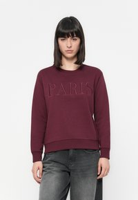 Mujer con cabello oscuro lacio, vistiendo una sudadera burdeos con "PARIS" bordado, combinada con jeans negros descoloridos, de pie frente a un fondo blanco.