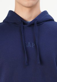 Felpa blu navy realizzata in morbido tessuto con cappuccio regolabile. Presenta il logo "GAP" in rilievo sul petto, creando un design pulito.