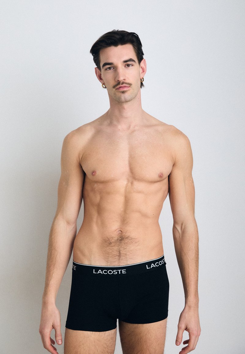 Boxer-briefs noirs pour hommes avec un logo Lacoste blanc sur la taille, ajustés et fabriqués dans un tissu lisse, portés par un mannequin masculin.