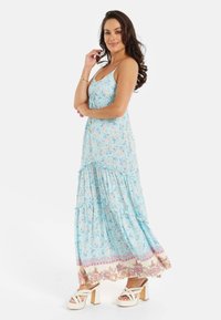 Light blue floral maxi dress with spaghetti straps, tiered ruffles, and patterned hem. Worn with cream chunky heeled sandals.