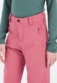 Růžové outdoorové kalhoty s kapsou na zip, plochým pasem na knoflík a hladkým povrchem. Spojené se zeleným topem.
