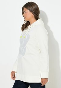 Sudadera de manga larga en color crema con un diseño gráfico en la parte frontal, capucha con cordones y aberturas laterales. Presenta detalles reflectantes y puños acanalados.