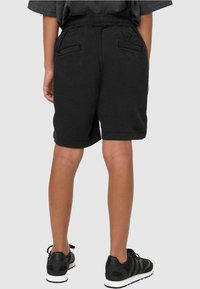 Shorts en coton noir avec une taille élastique, comportant des poches arrière et des ourlets roulés. Portés avec des chaussures de sport noires ayant des semelles blanches.