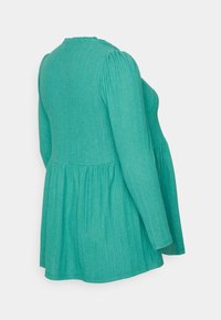 Top verde malhado com mangas longas, tecido texturizado e silhueta peplum. Apresenta um decote redondo e costuras de ombro franzidas.